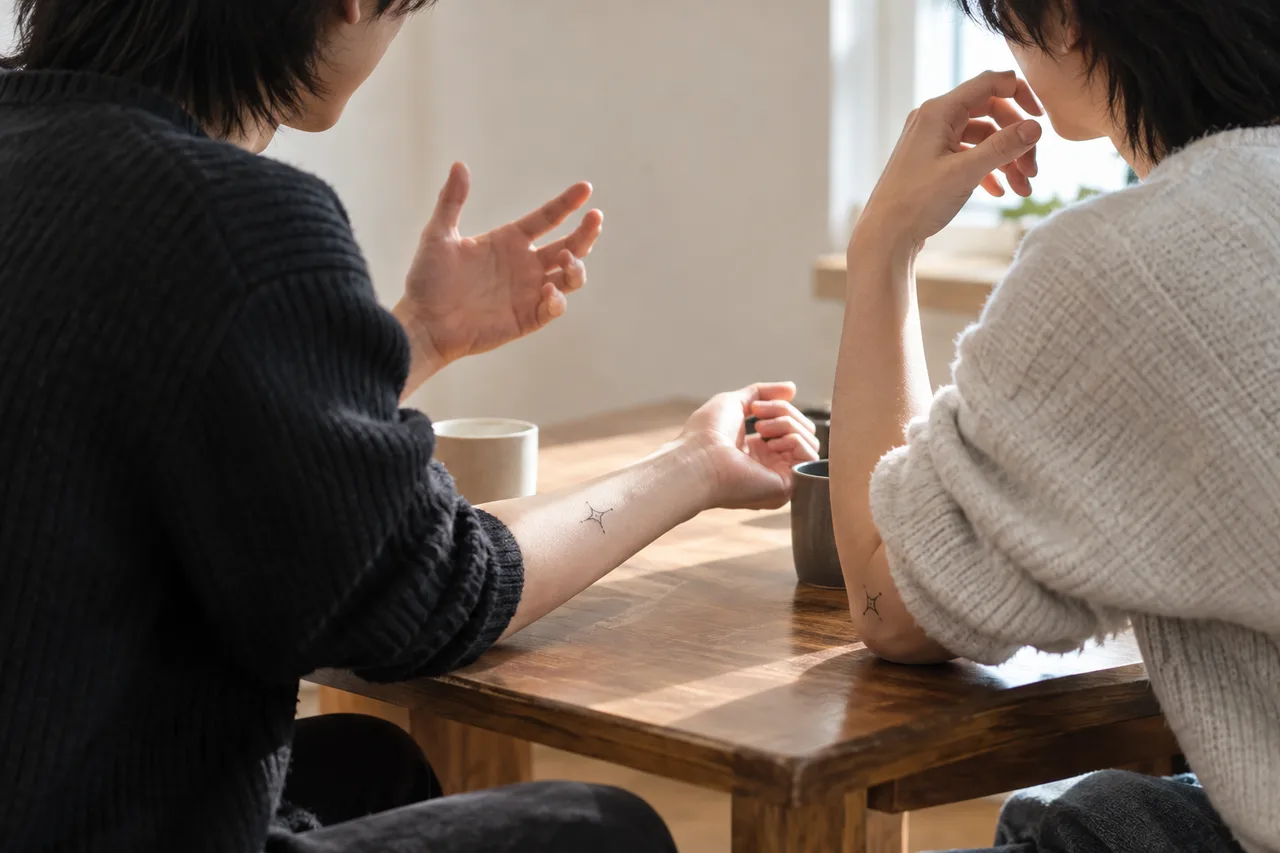 뒷모습의 성인 두 명이 나무 탁자에서 대화하며, 팔에 작은 별 모양의 비슷한 타투가 보인다. 탁자에는 컵이 놓여 있다.